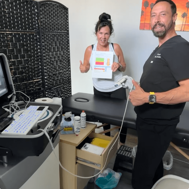 Medical professional giving thumbs up beside a woman seated on an exam table holding a bone density report in a medical exam room with ultrasound equipment.
