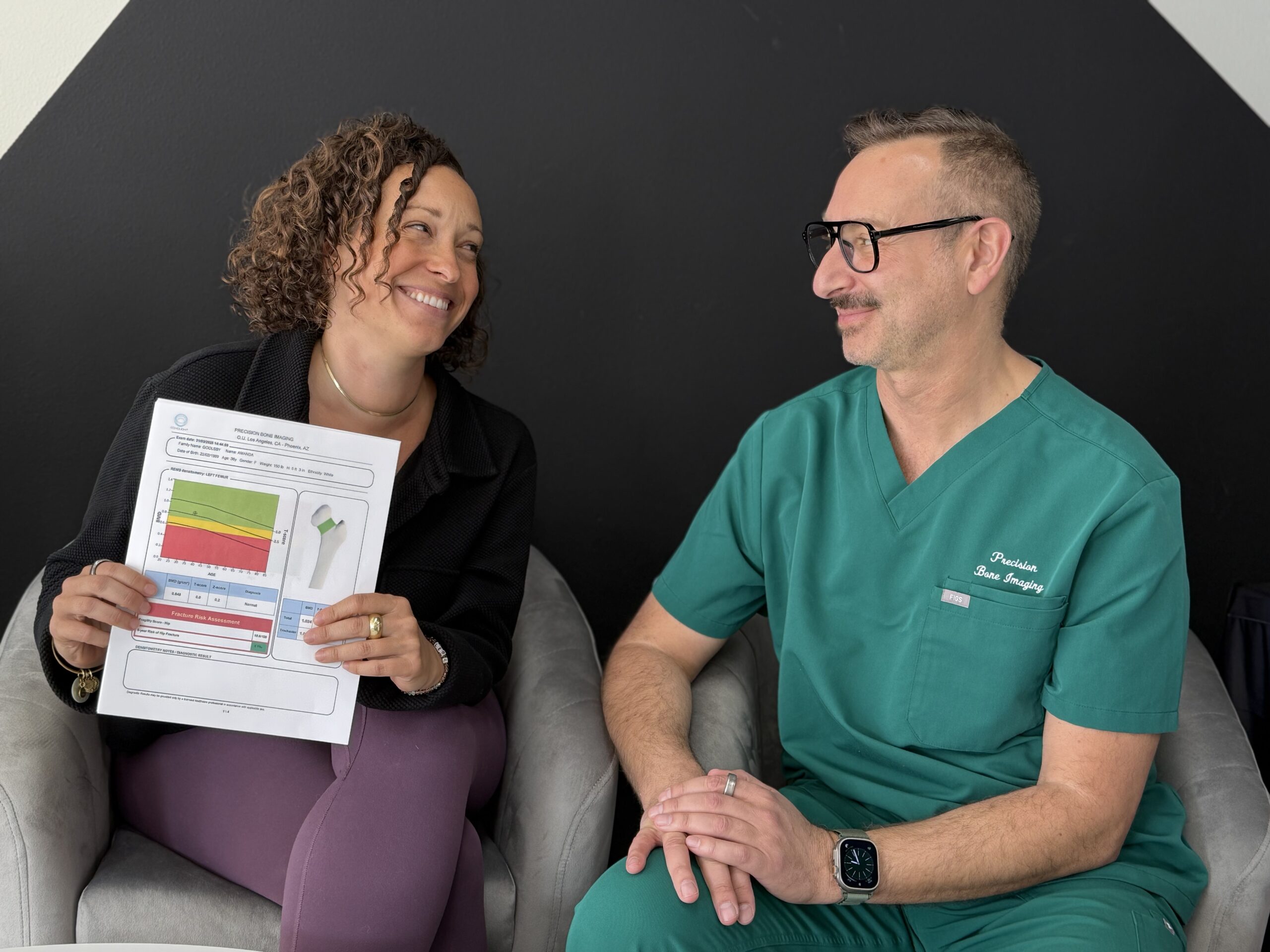 Smiling woman with curly hair holds a color-coded medical report while sitting next to a man in teal medical scrubs; they are looking at each other in front of a dark wall.