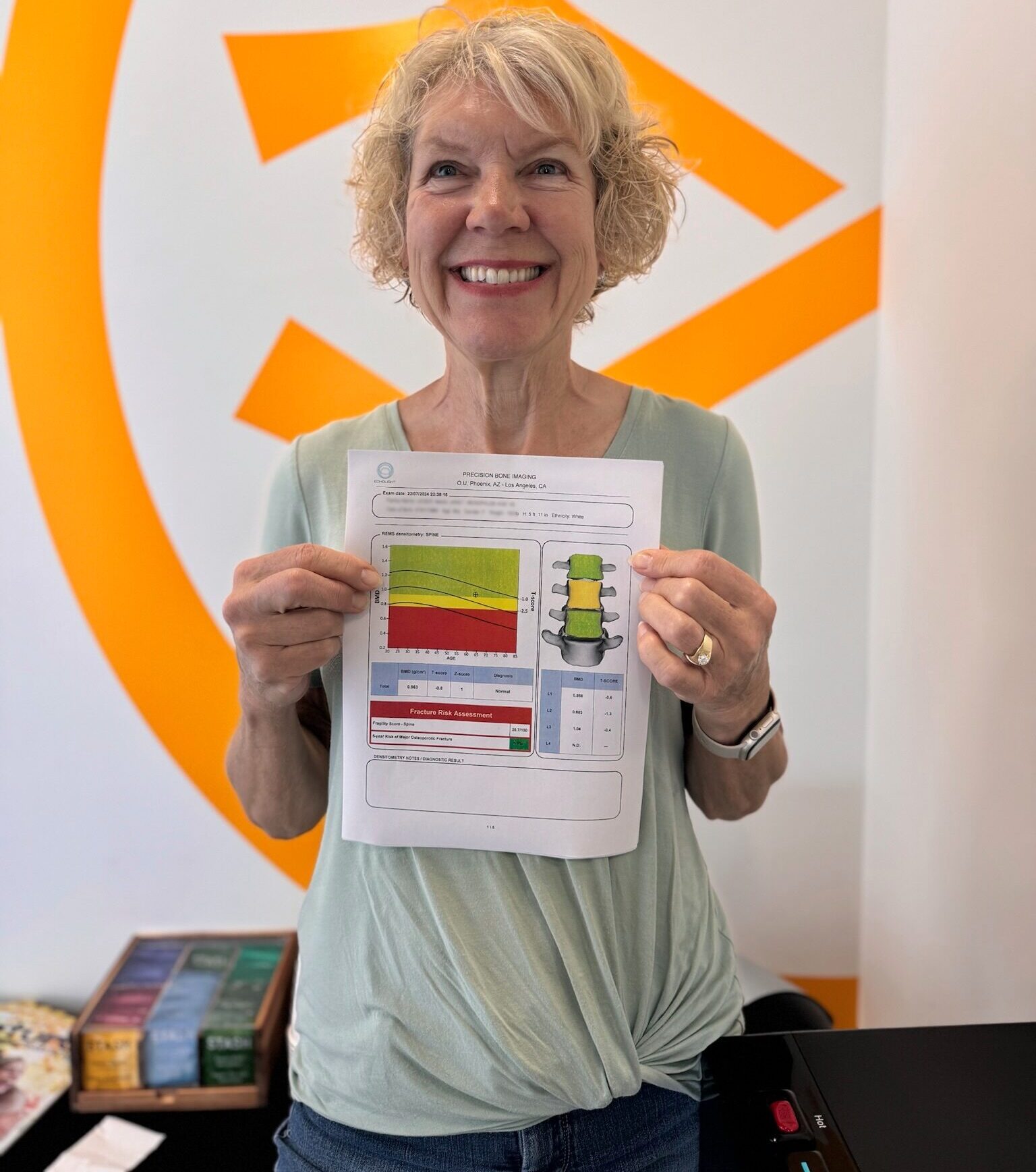 Smiling woman with short curly hair holding a bone density report in front of a large orange design on a wall.