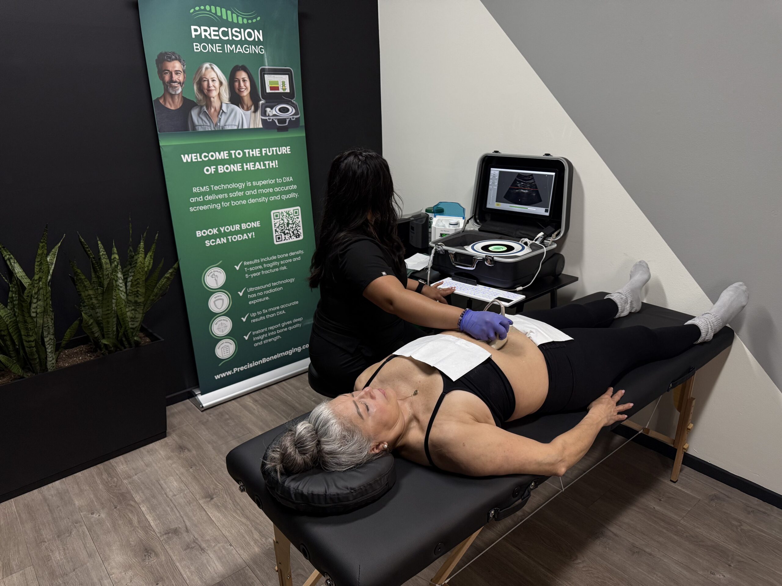Technician wearing gloves performs a bone density scan on a woman lying on an exam table in a clinic, with a Precision Bone Imaging banner in the background.
