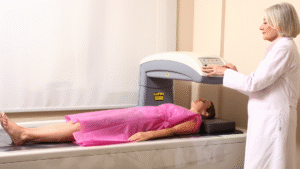 A patient, wearing a pink gown, lies on a table, receiving a Bone Density Scan (DXA/DEXA) conducted by a female healthcare professional in a white coat.