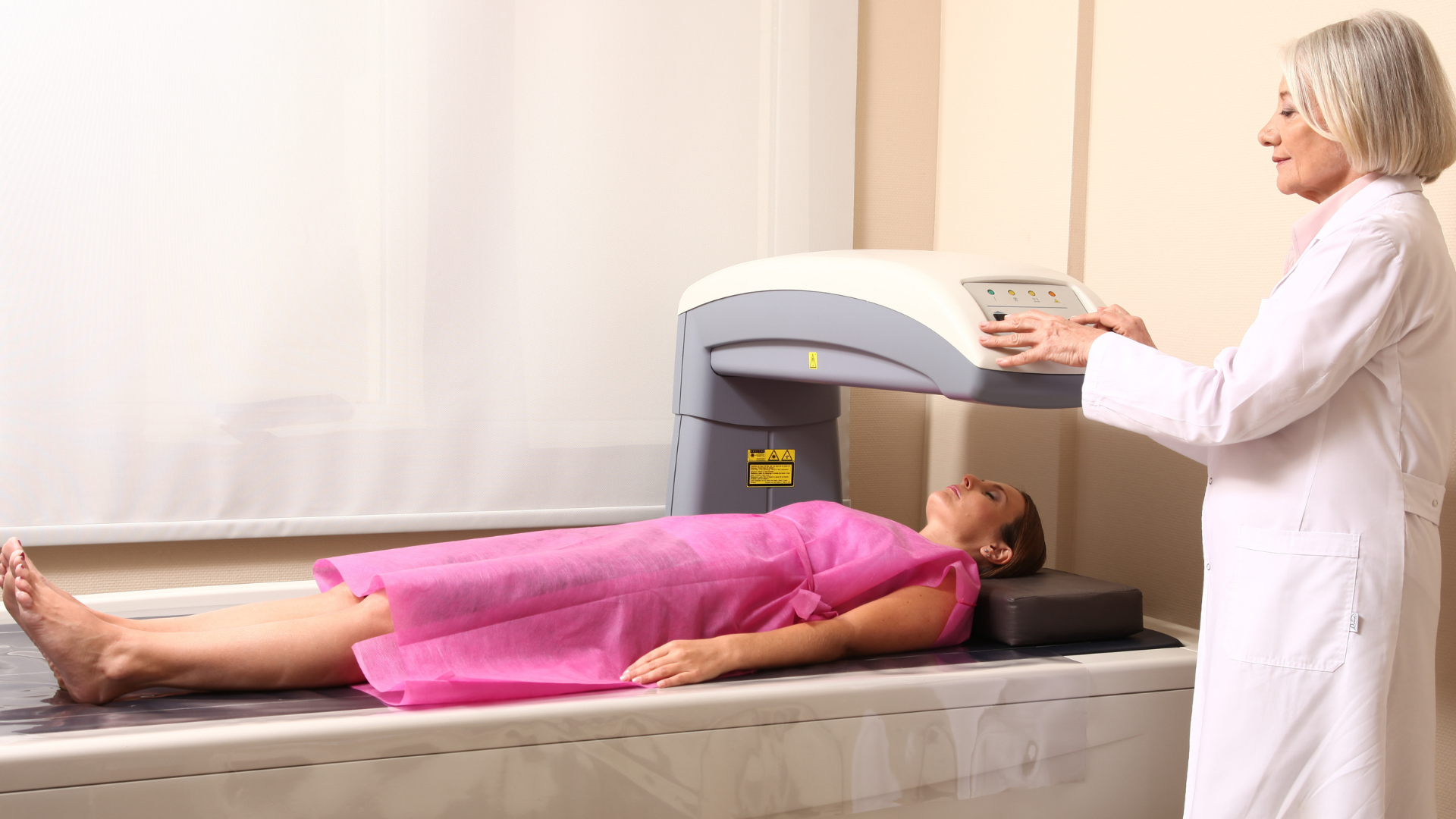 A patient, wearing a pink gown, lies on a table, receiving a Bone Density Scan (DXA/DEXA) conducted by a female healthcare professional in a white coat.
