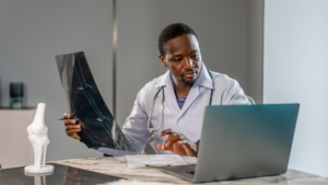 Doctor reviewing bone density X-ray while working at desk with laptop; bone density timeline guide concept.