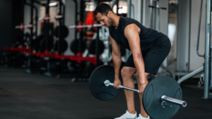 Man performing deadlift with barbell in gym: focused athlete in black tank top and shorts lifting heavy weight from the floor, surrounded by weight racks and equipment. Precision Bone Imaging – highlighting exercise-driven bone growth.